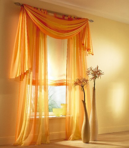 Aranżacja okienna z pomarańczowych, zwiewnych firan i rolet rzymskich, dopełniona wysokimi, smukłymi wazonami z dekoracyjnymi gałązkami.