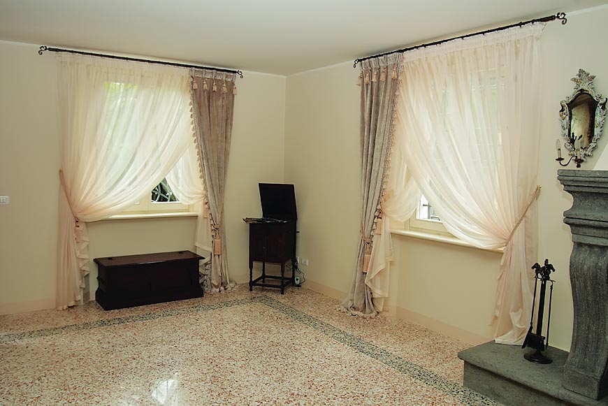 Jasne wnętrze z dwoma oknami, udekorowanymi beżowo-białymi, drapowanymi firanami z ozdobnymi chwostami, ciemny kredens między oknami, marmurowa podłoga w terazzo i fragment kamiennego kominka...