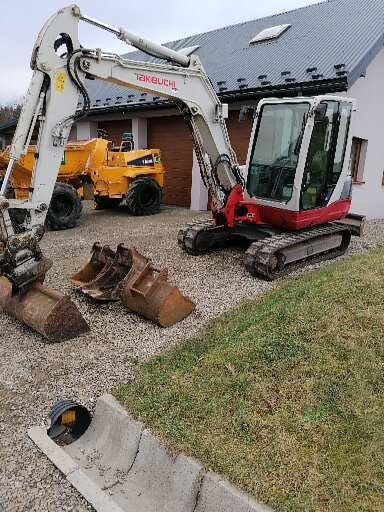 Mała koparka Takeuchi z łyżkami na żwirowym podłożu, w tle żółty walec i dom z szarą dachówką.