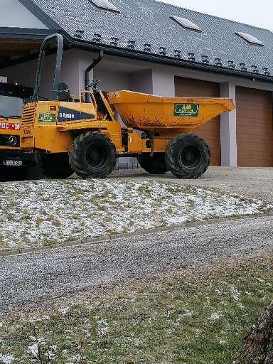 Żółta wywrotka budowlana stoi na placu budowy pokrytym śniegiem, z widocznym budynkiem w tle.