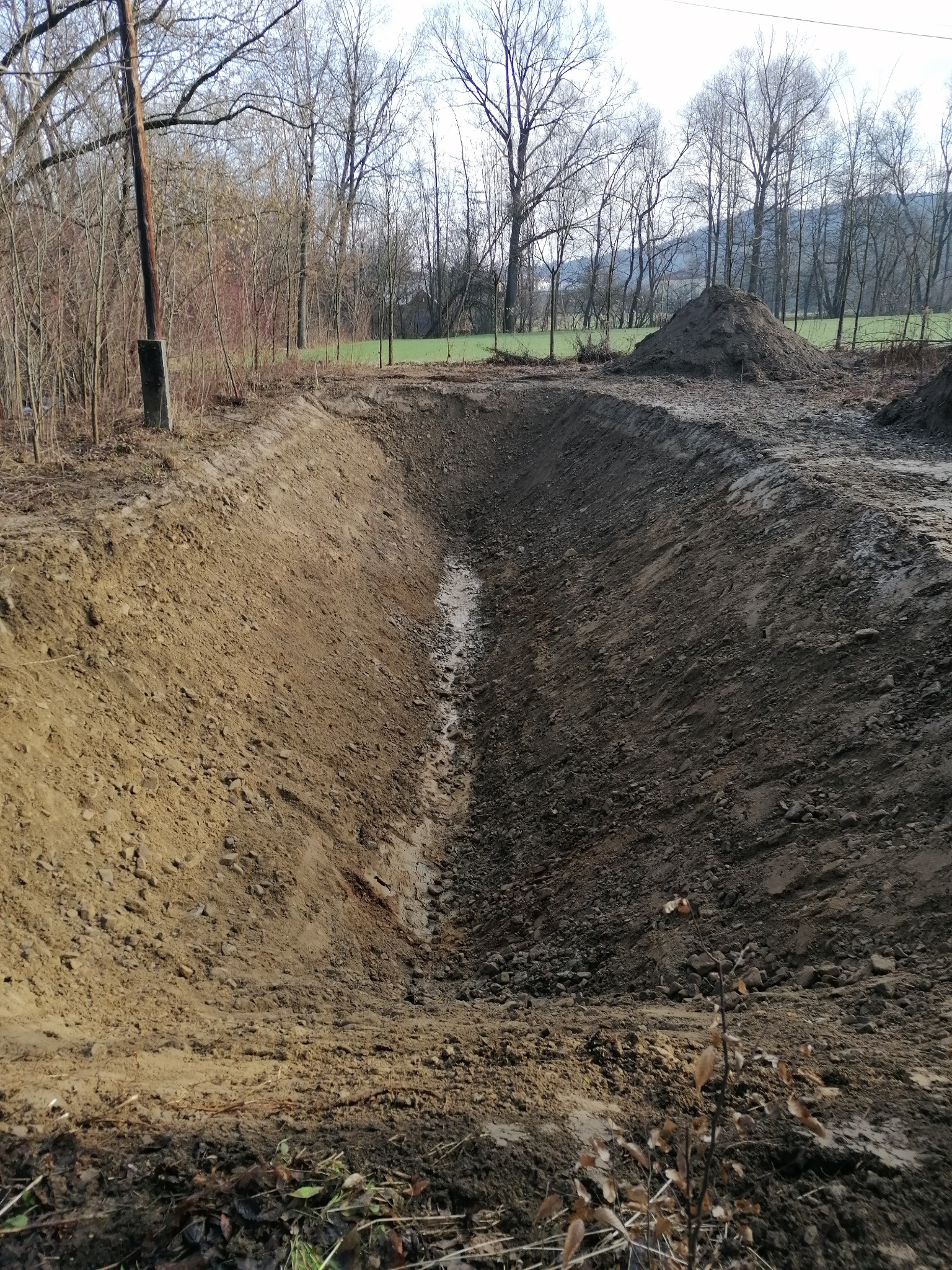 Długi, wykopany rów w ziemi, widoczny stos ziemi w tle, krajobraz wiejski z drzewami bez liści.