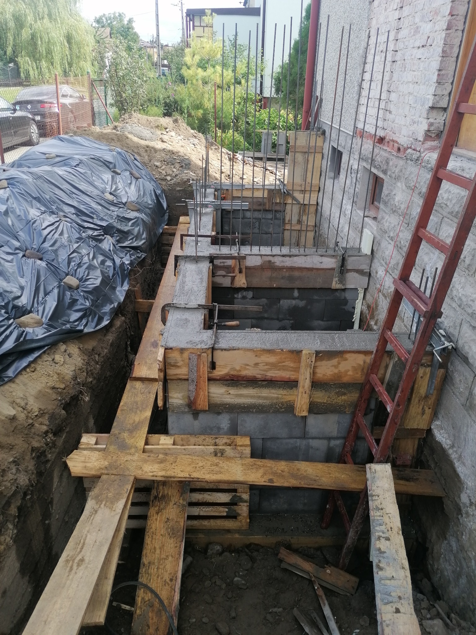 Wykop fundamentowy z częściowo wylanym betonem, wzmocniony deskowaniem i zbrojeniem, przy zewnętrznej ścianie budynku z cegły, z widoczną drabiną i czarną folią zabezpieczającą.