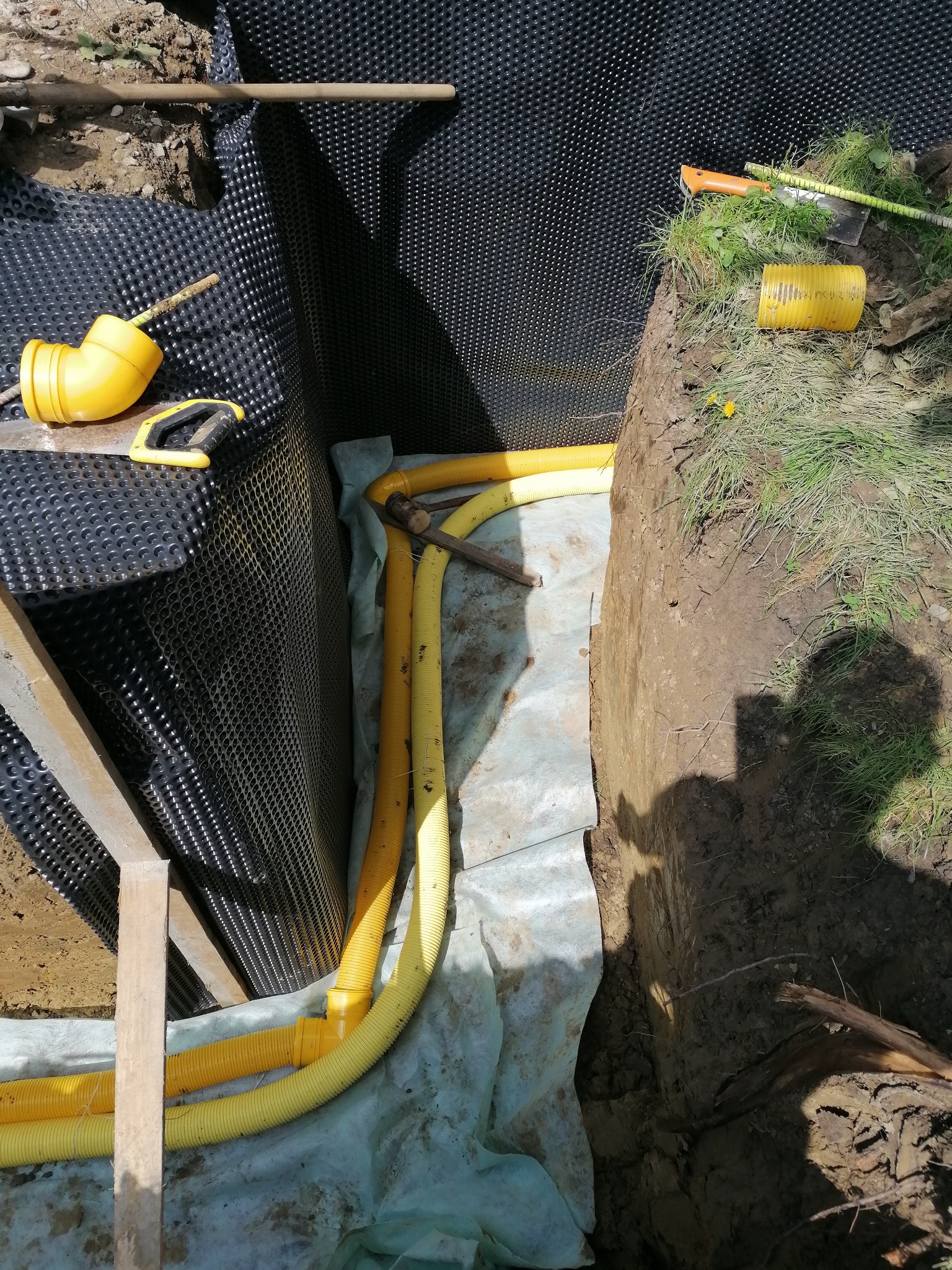 Żółte rury drenarskie ułożone w wykopie wyłożonym geowłókniną i folią kubełkową, widoczne narzędzia i ziemia.