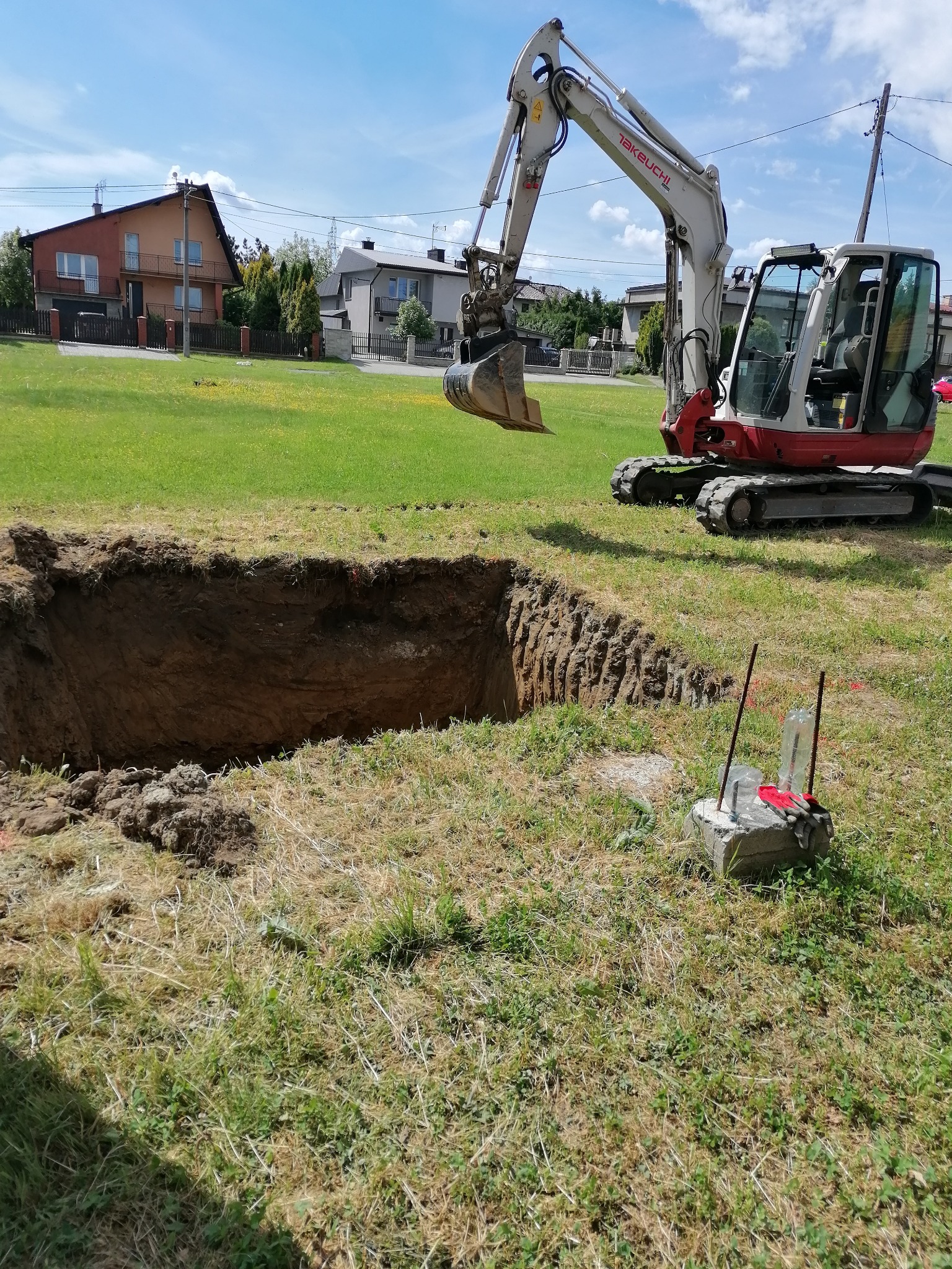 Świeżo wykopany dół w ziemi, prawdopodobnie pod fundamenty, obok stoi biało-czerwony minikoparka Takeuchi oraz betonowy słupek z wbitymi prętami i rękawicami roboczymi na wierzchu, w tle zabudowa...