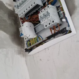 Otwarta, biała skrzynka rozdzielcza z widocznymi bezpiecznikami, przewodami w różnych kolorach i listwami zaciskowymi zamontowana na surowej, szarej ścianie. Widok od dołu, ukazujący szczegóły...