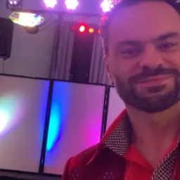 Uśmiechnięty mężczyzna w czerwonej koszuli na tle sceny z oświetleniem imprezowym, widoczne reflektory i panele LED zmieniające kolory.