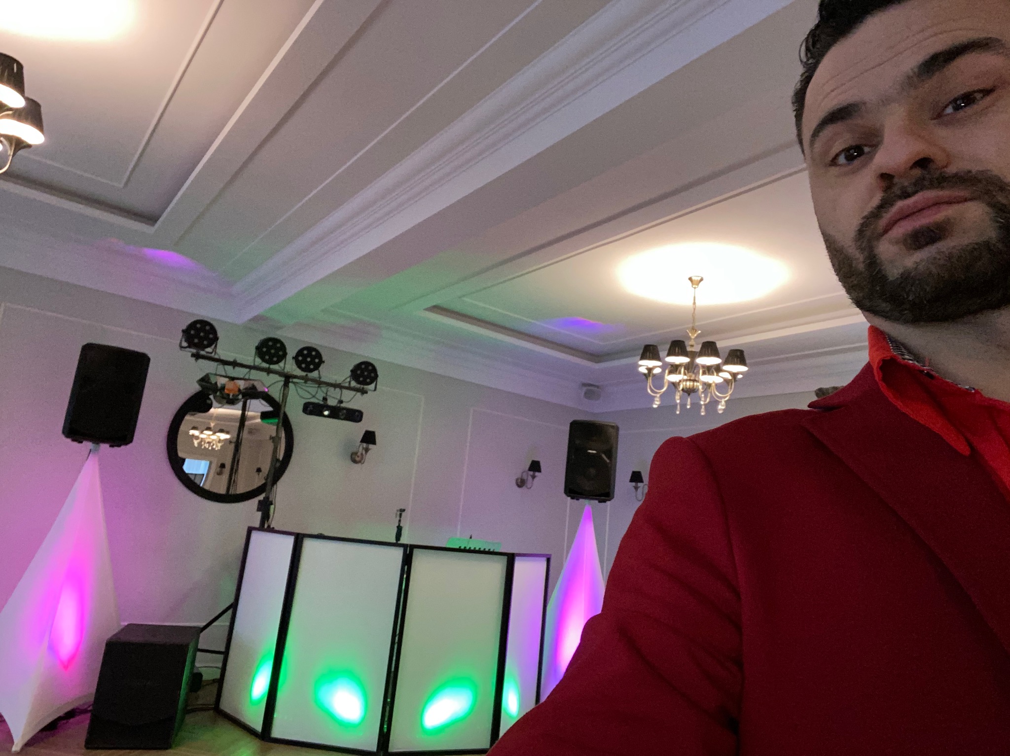 Mężczyzna w czerwonej marynarce pozuje na tle sprzętu DJ-skiego z podświetleniem LED na sali z żyrandolem.