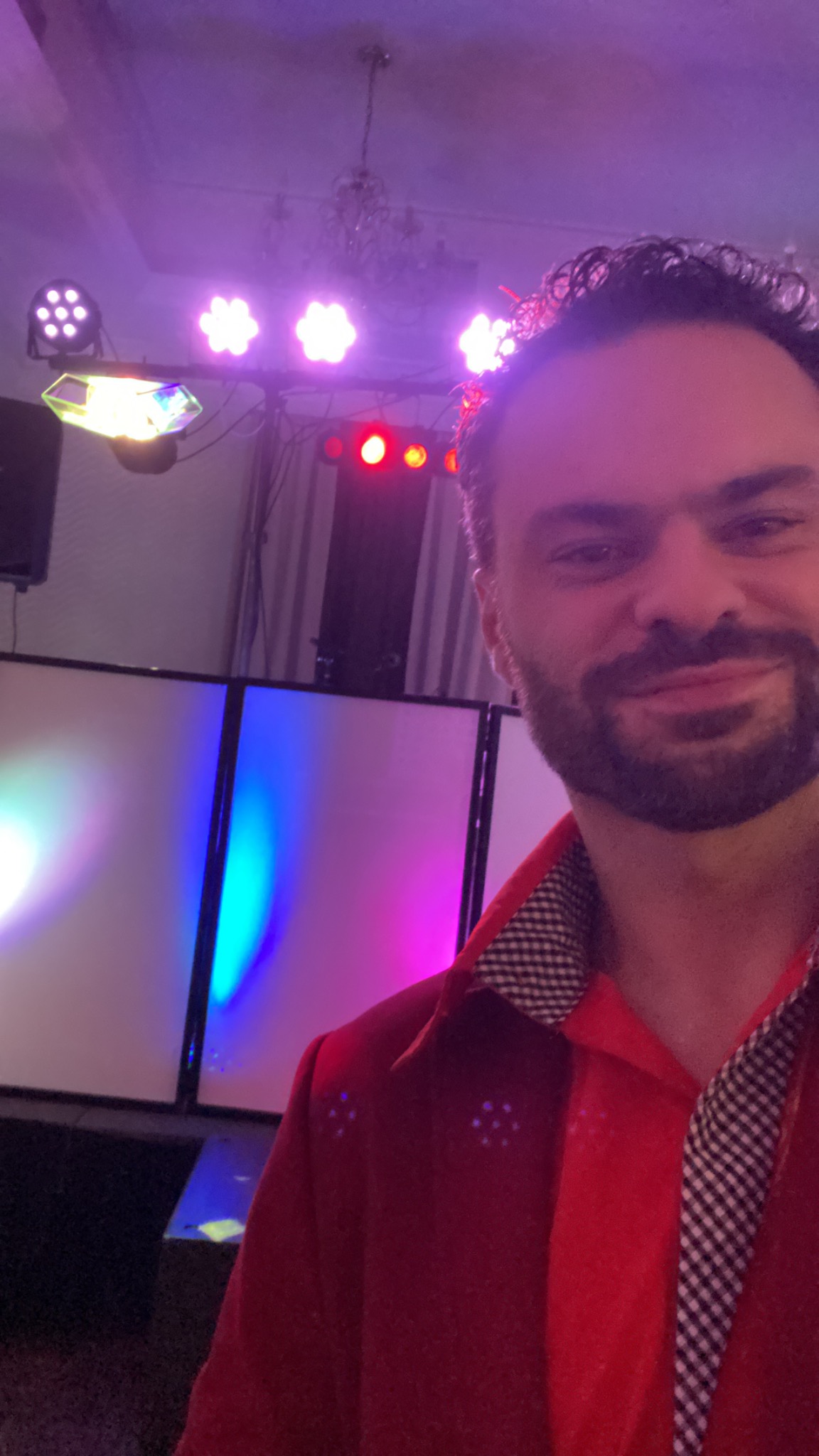 Uśmiechnięty mężczyzna w czerwonej koszuli na tle sceny z oświetleniem imprezowym, widoczne reflektory i panele LED zmieniające kolory.