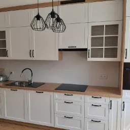 Nowoczesna kuchnia z białymi szafkami i drewnianymi blatami, oświetlona trzema czarnymi lampami wiszącymi o geometrycznych kloszach. Widoczne czarne uchwyty szafek, zlewozmywak, płyta indukcyjna...