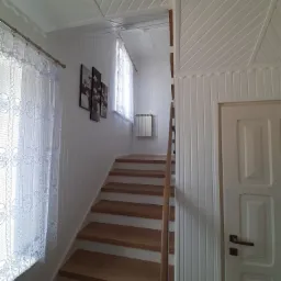 Klatka schodowa w jasnym wnętrzu z drewnianymi schodami, białymi ścianami z boazerią i oknami z koronkowymi firankami. Na ścianie wiszą cztery obrazy z motywem kwiatowym.