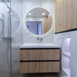 Nowoczesna łazienka z kabiną prysznicową ze szklaną ścianką, okrągłym lustrem z podświetleniem LED, drewnianą szafką pod umywalką z czarnymi uchwytami i zabudową meblową z drewna.