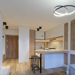 MyBigBoxArchitekci Sp z o.o. - Nowoczesna kuchnia z wyspą i hokerami w mieszkaniu w Warszawie, oświetlona wiszącą lampą z okręgów, z elementami drewna i bieli.