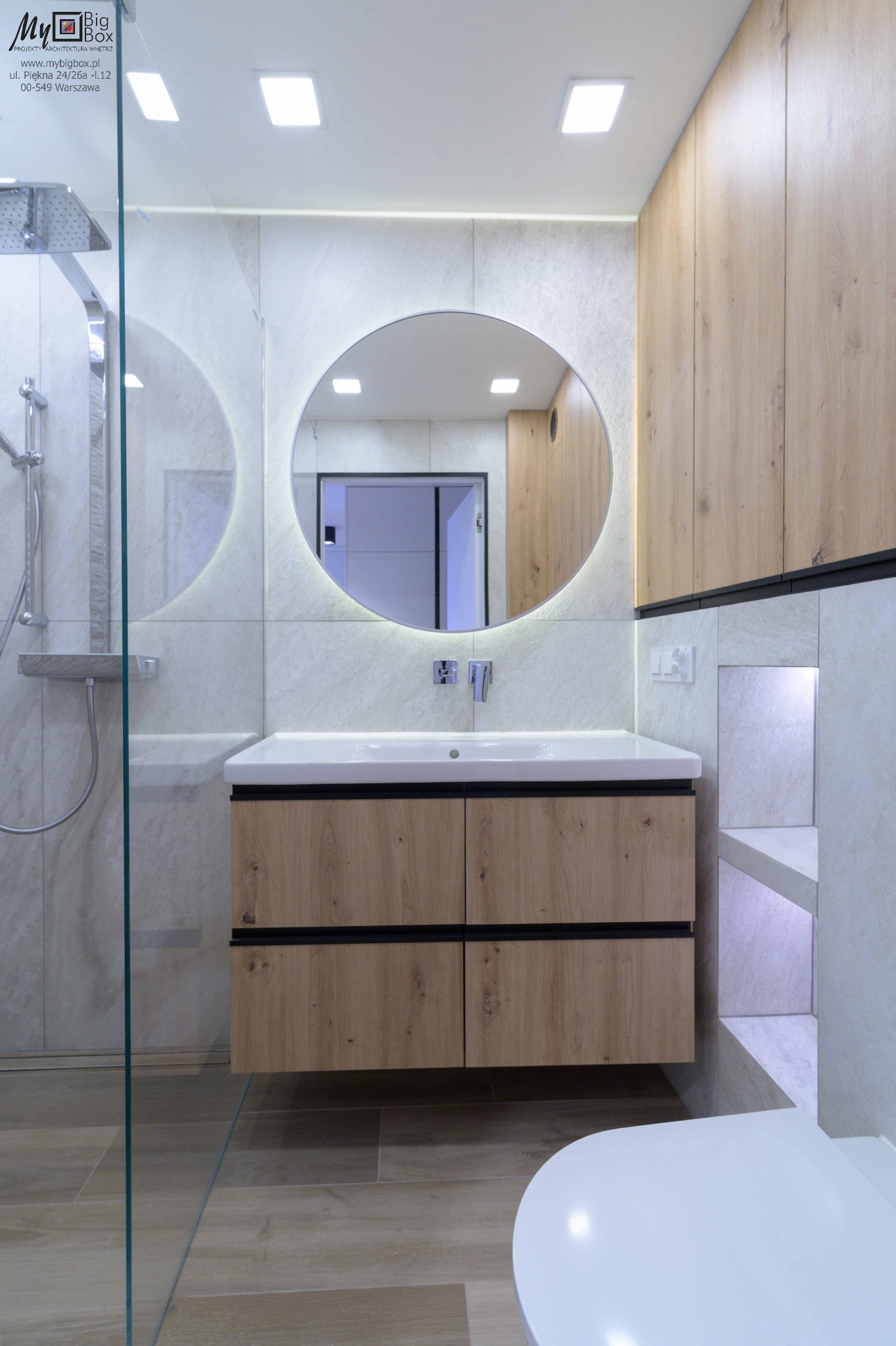 Nowoczesna łazienka z kabiną prysznicową ze szklaną ścianką, okrągłym lustrem z podświetleniem LED, drewnianą szafką pod umywalką z czarnymi uchwytami i zabudową meblową z drewna.