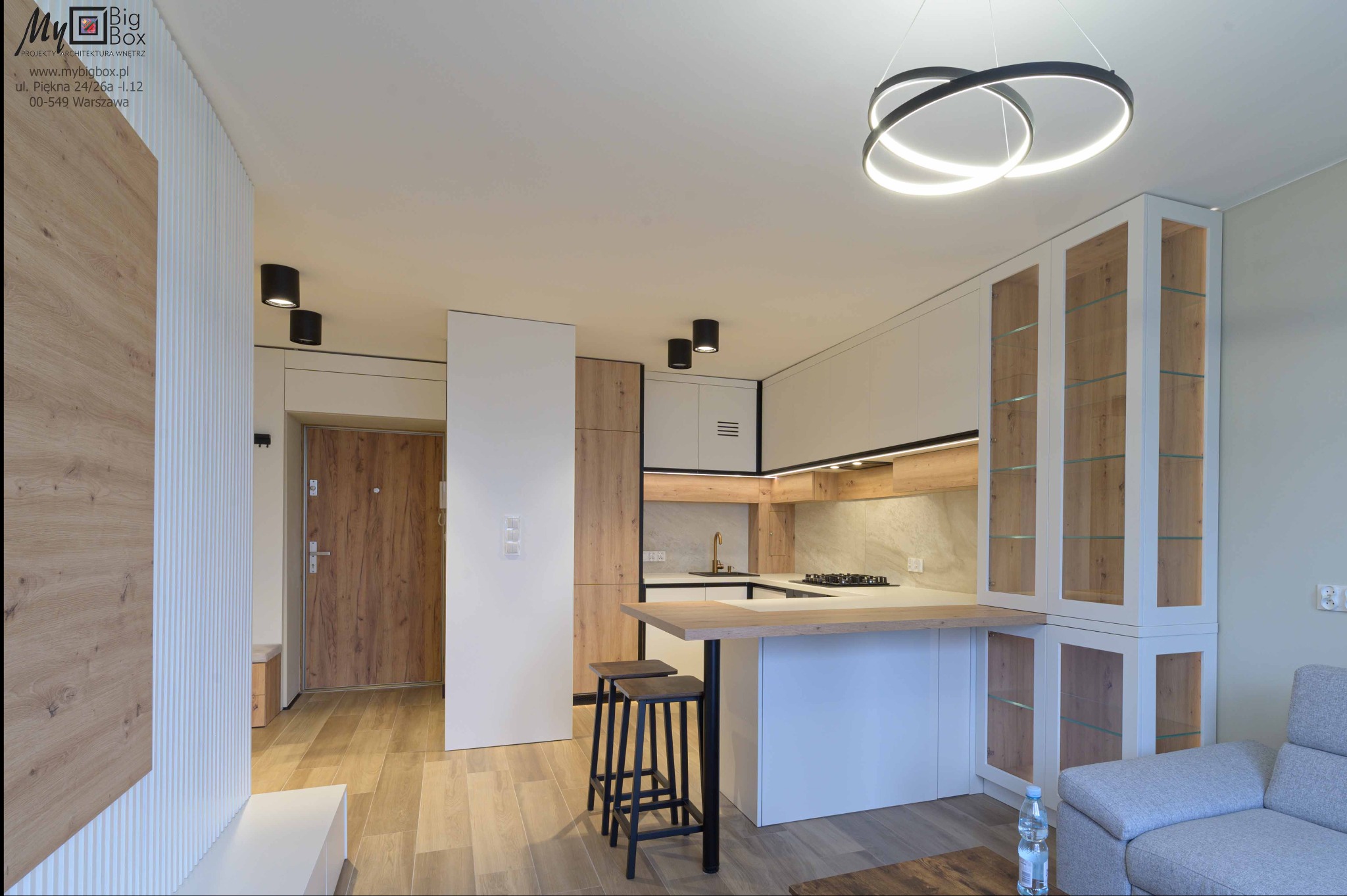 Nowoczesna kuchnia z wyspą i hokerami w mieszkaniu w Warszawie, oświetlona wiszącą lampą z okręgów, z elementami drewna i bieli.