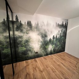 Michał Brzyski - Pokój po remoncie: drewniana podłoga, ściana z fototapetą leśną. Widoczne gniazdka elektryczne i fragment szafy z czarną ramą. Minimalistyczny wystrój wnętrza.