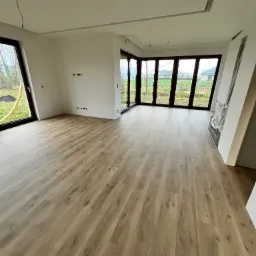 Jasne panele podłogowe ułożone w przestronnym pokoju z dużymi oknami w Biłgoraju. Ściany pomalowane na biało, widoczne gniazdka elektryczne.