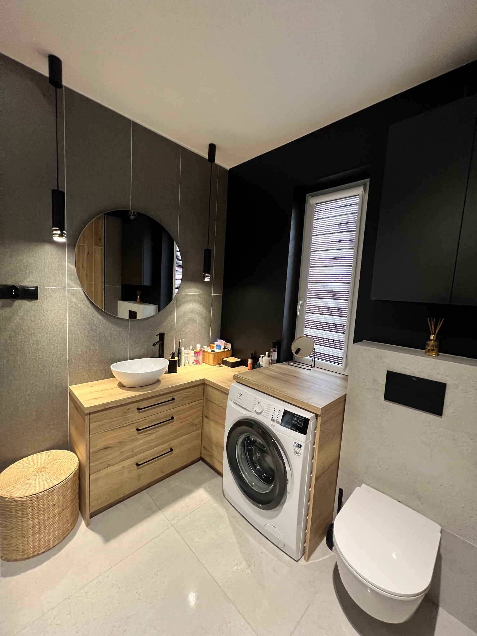 Nowoczesna łazienka z drewnianą zabudową umywalki, okrągłym lustrem, pralką wkomponowaną w blat i wiszącą toaletą. Minimalistyczne oświetlenie punktowe.