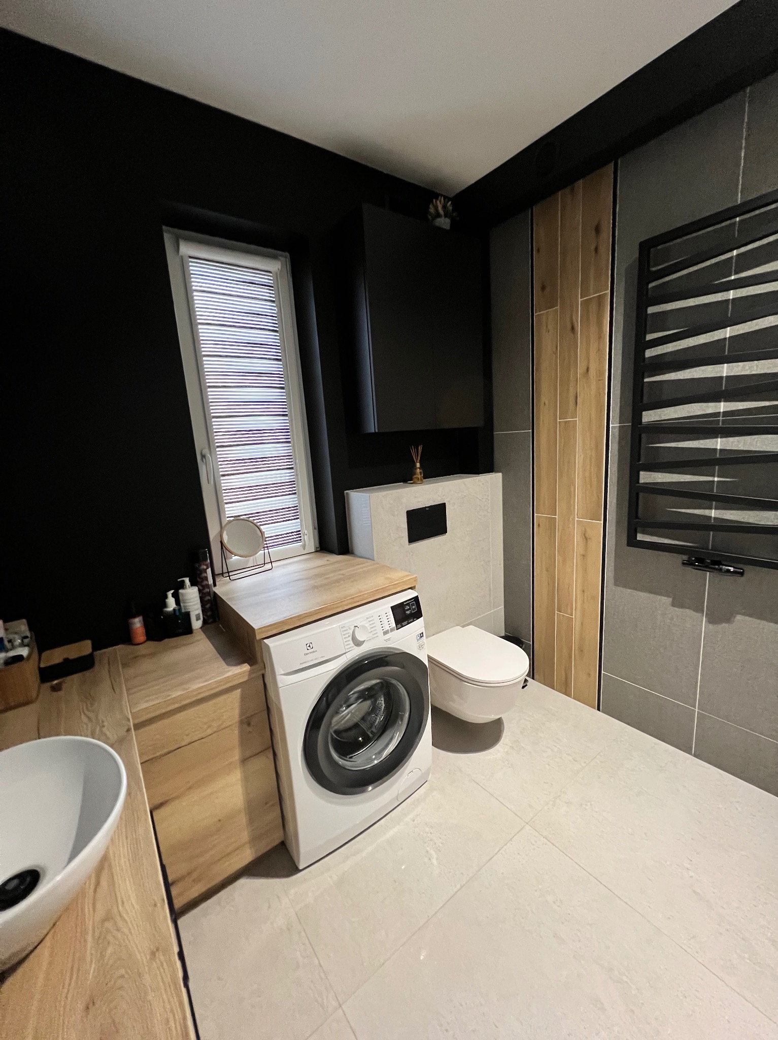 Nowoczesna łazienka w ciemnych kolorach z drewnianymi akcentami. Uwagę przykuwa pralka wkomponowana w drewniany blat, obok umywalka i toaleta.