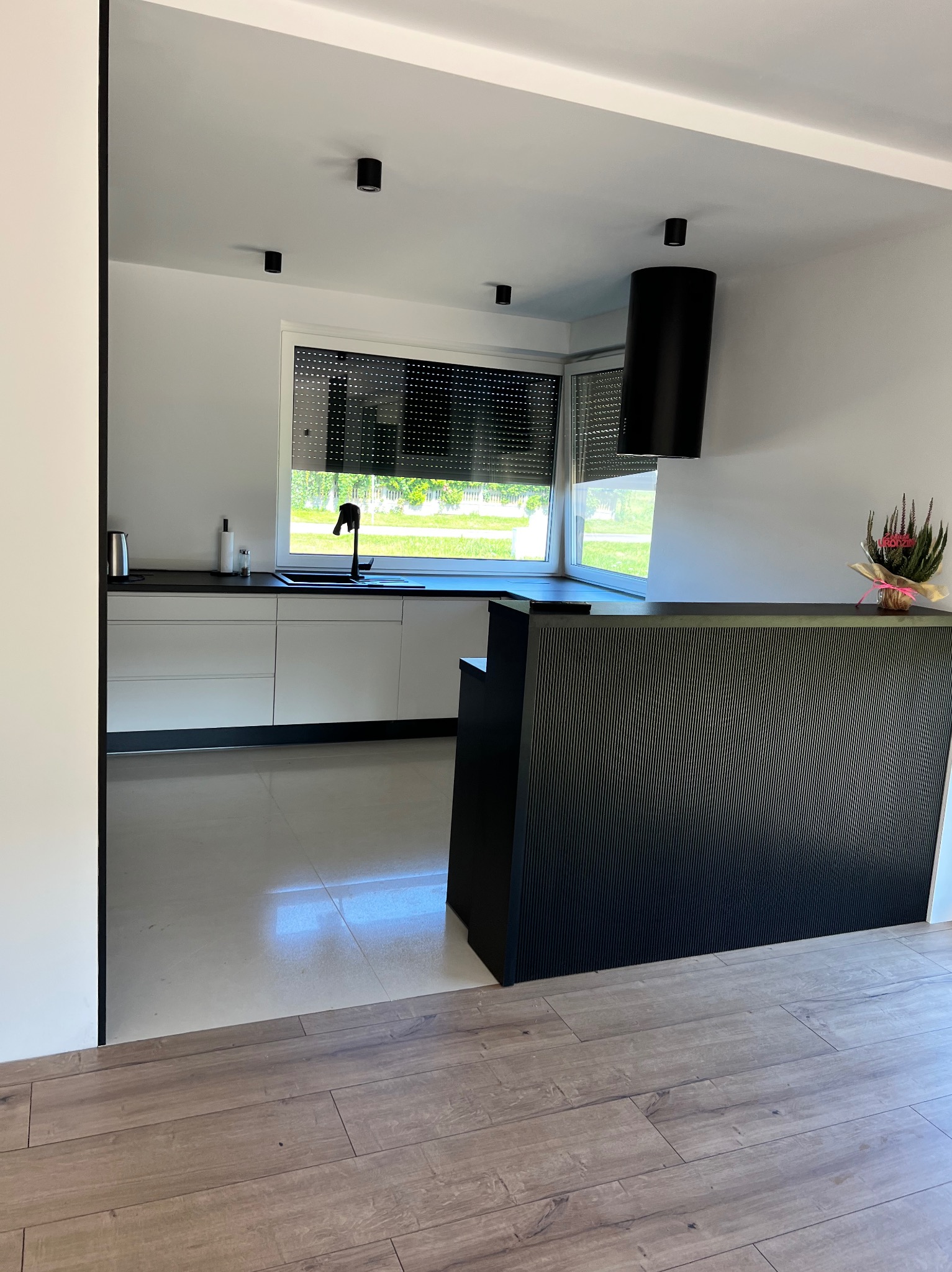 Nowoczesna kuchnia w bieli i czerni z oknem i minimalistycznym okapem cylindrycznym. Elementy dekoracyjne, takie jak kwiaty, dodają subtelności wnętrzu.