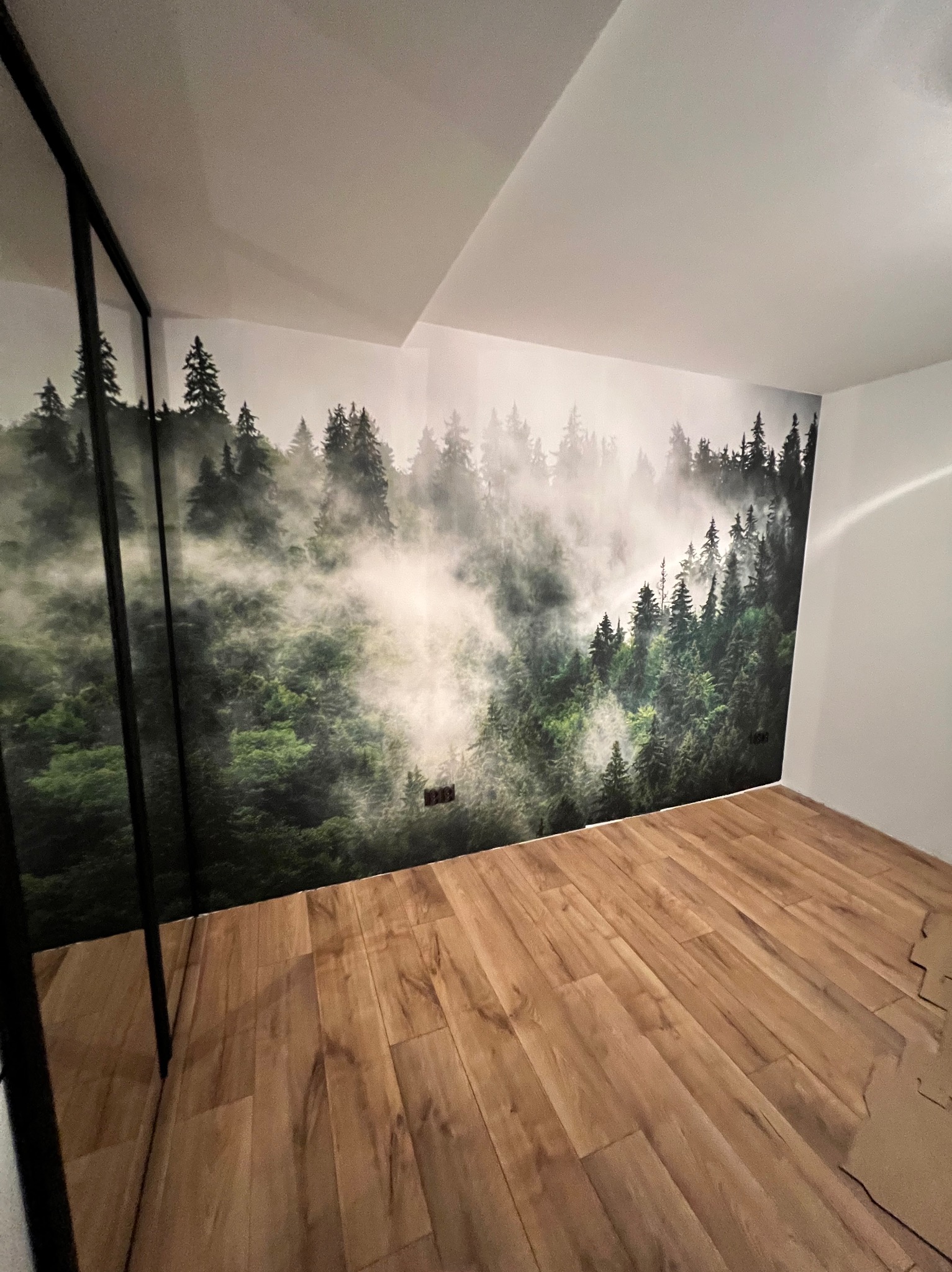 Pokój po remoncie: drewniana podłoga, ściana z fototapetą leśną. Widoczne gniazdka elektryczne i fragment szafy z czarną ramą. Minimalistyczny wystrój wnętrza.