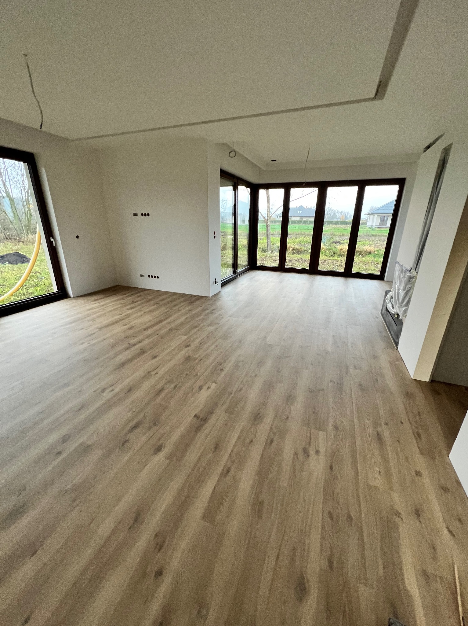 Jasne panele podłogowe ułożone w przestronnym pokoju z dużymi oknami w Biłgoraju. Ściany pomalowane na biało, widoczne gniazdka elektryczne.