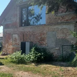 Ceglana ściana budynku z nowym, dużym oknem, częściowo otynkowana, z widocznymi śladami napraw i roślinnością u podstawy. Na pierwszym planie trawnik z fragmentami ziemi.