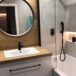 Nowoczesna łazienka z drewnianą okładziną, okrągłym lustrem, białą umywalką nablatową z czarną armaturą, kabiną prysznicową typu walk-in z czarnymi elementami i mozaikową podłogą.