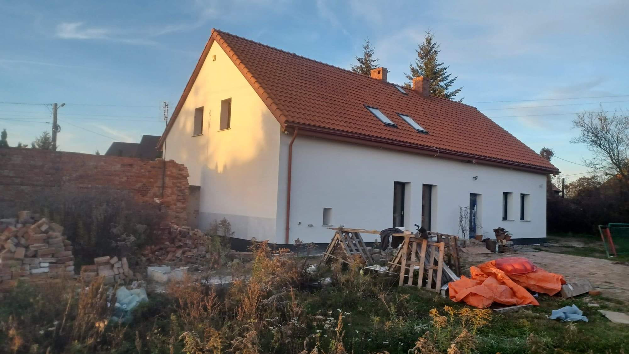 Dom jednorodzinny po renowacji elewacji, widoczne świeżo pomalowane ściany w odcieniach bieli, czerwona dachówka, gruz ceglany i materiały budowlane na pierwszym planie.