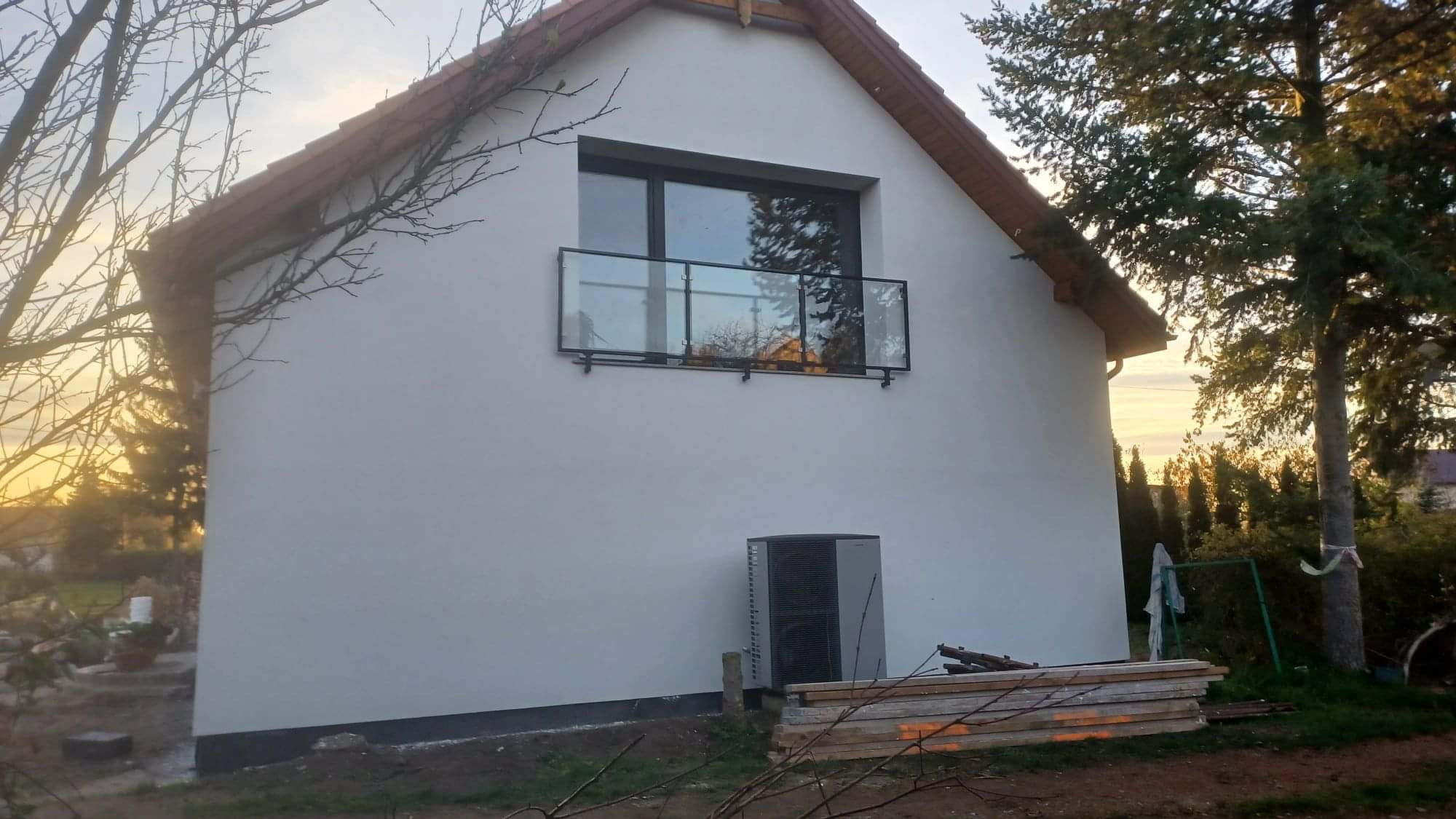 Biała ściana domu jednorodzinnego we Wrocławiu, z oknem balkonowym z szklaną balustradą, klimatyzatorem na zewnątrz i stertą desek u podnóża ściany.