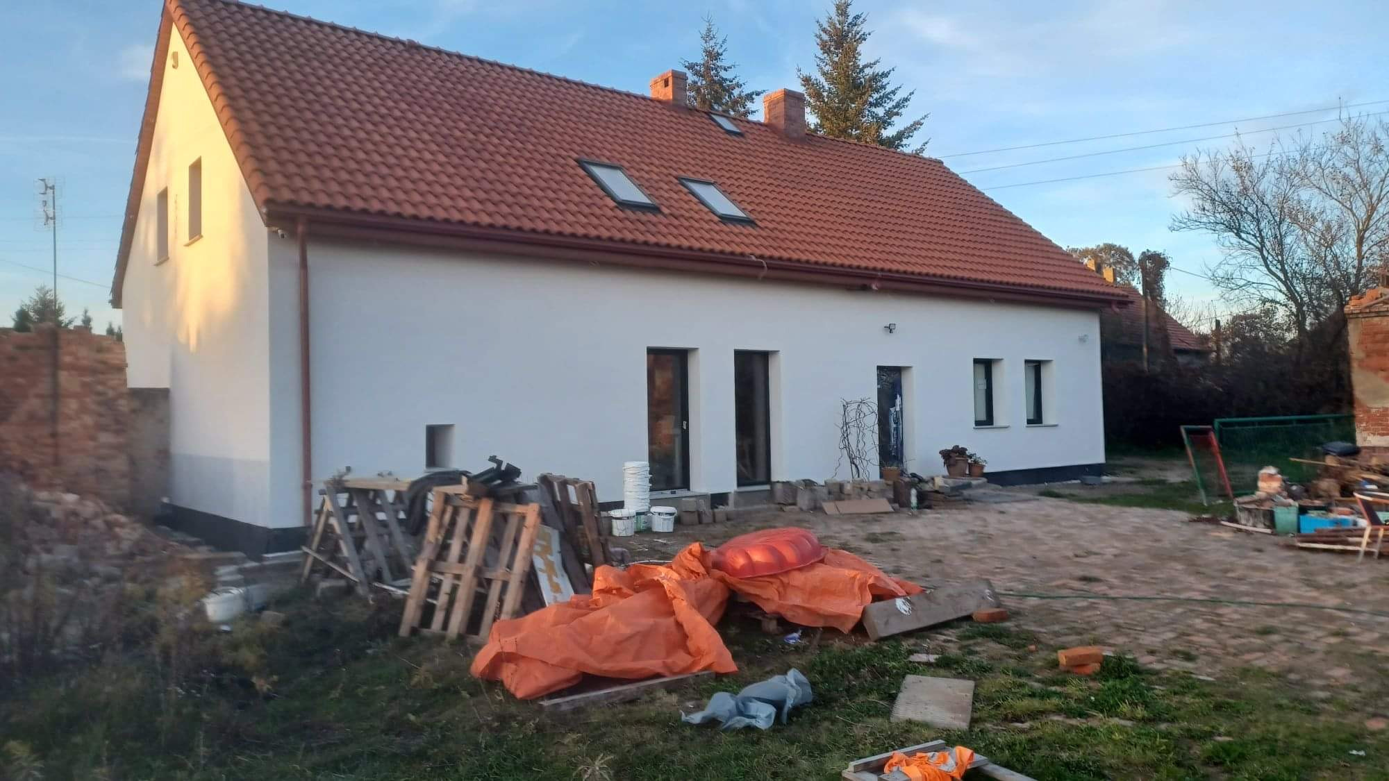 Świeżo pomalowany dom jednorodzinny z czerwoną dachówką, widoczne palety, wiaderka po farbie i pomarańczowe plandeki na posesji w trakcie prac wykończeniowych.