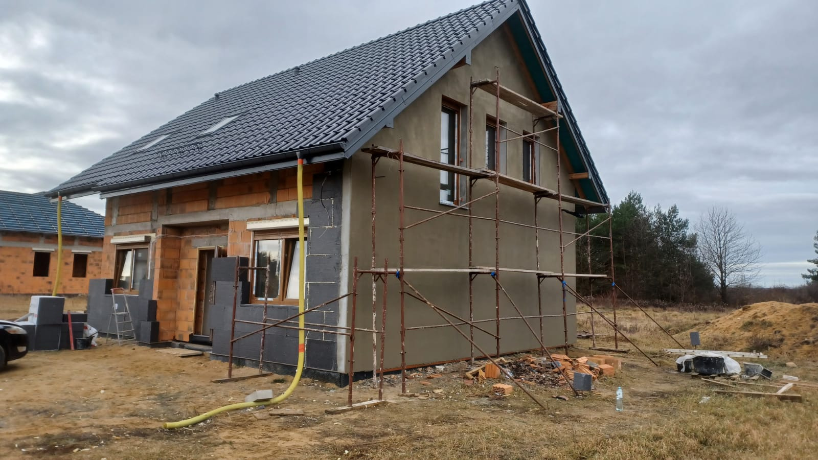 Dom jednorodzinny w trakcie ocieplania ścian zewnętrznych, widoczne rusztowania, częściowo otynkowana elewacja, styropian i żółty wąż do podawania materiałów.