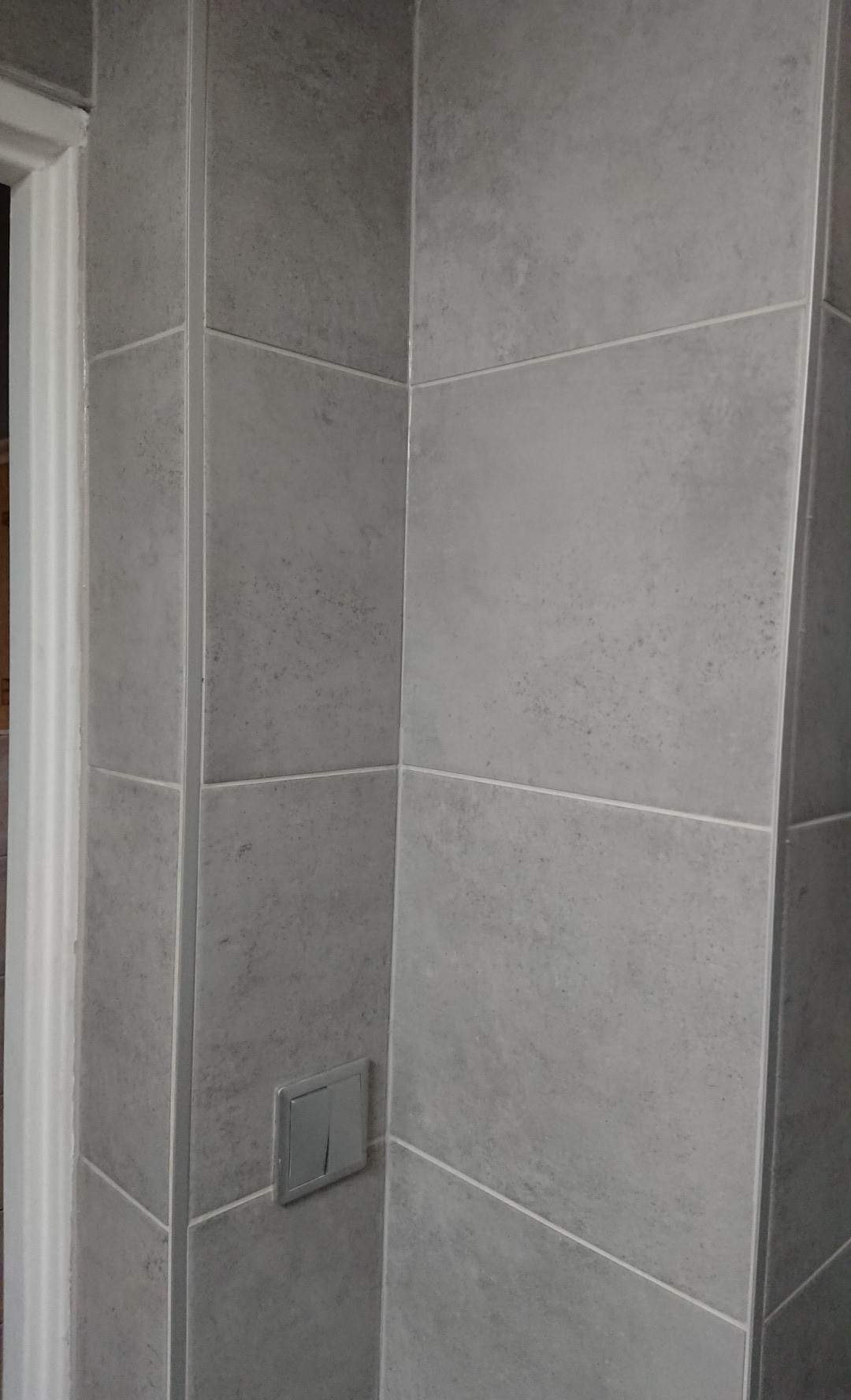 Wykończona ściana w łazience z szarymi, kwadratowymi płytkami i chromowanym włącznikiem światła, widoczne fugi i fragment futryny drzwi.