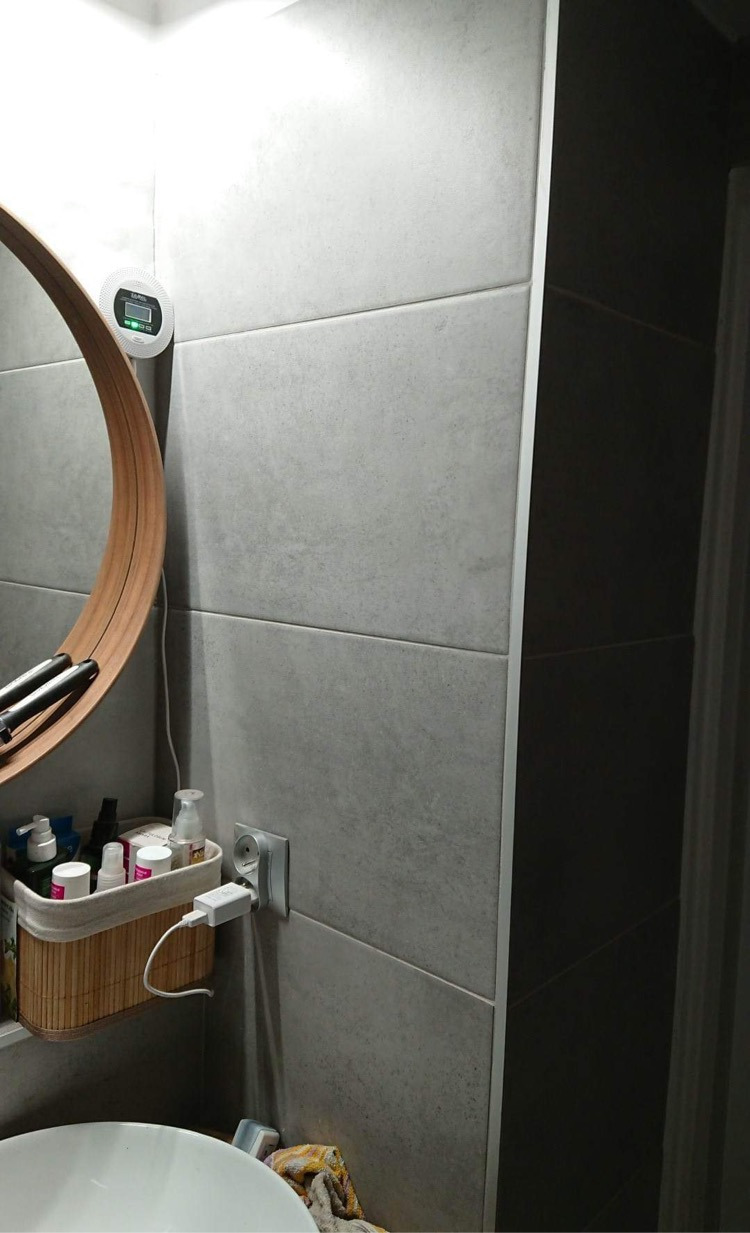 Fragment wykończonej łazienki z szarymi, matowymi płytkami, okrągłym lustrem z drewnianą ramą, koszykiem wiklinowym na kosmetyki i czujnikiem czadu na ścianie.