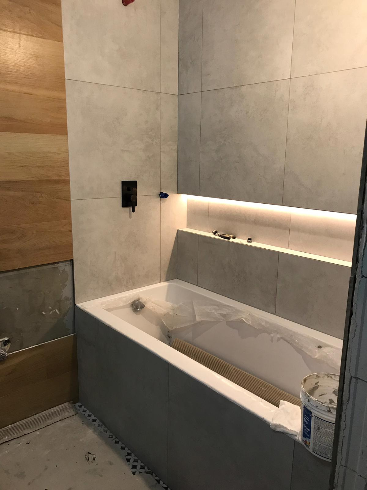 Łazienka w trakcie wykańczania: szare płytki ścienne, zabudowana wanna z zabezpieczeniem, drewniana okładzina ściany, widoczne narzędzia i materiały budowlane.