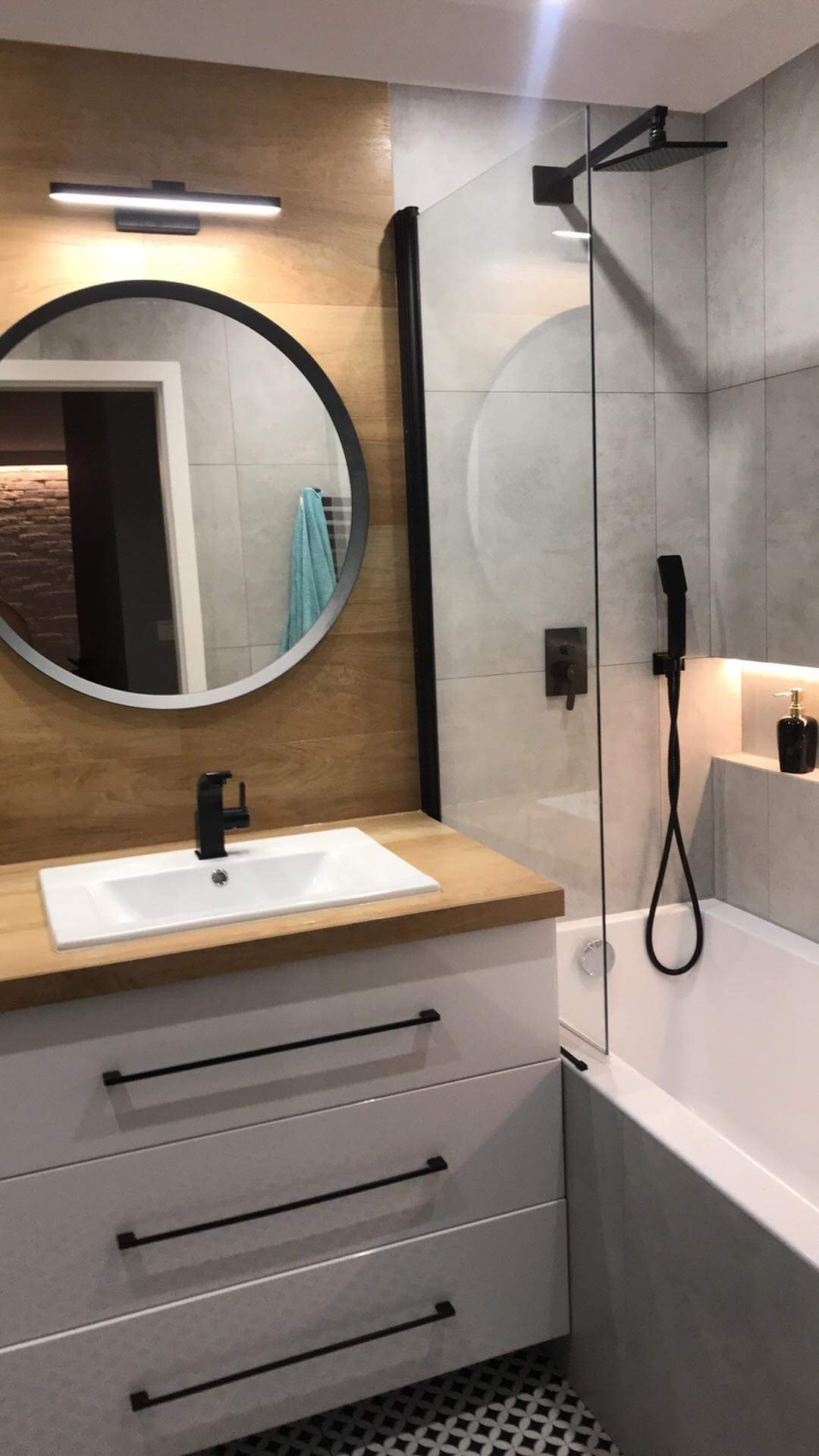 Nowoczesna łazienka z drewnianą okładziną, okrągłym lustrem, białą umywalką nablatową z czarną armaturą, kabiną prysznicową typu walk-in z czarnymi elementami i mozaikową podłogą.