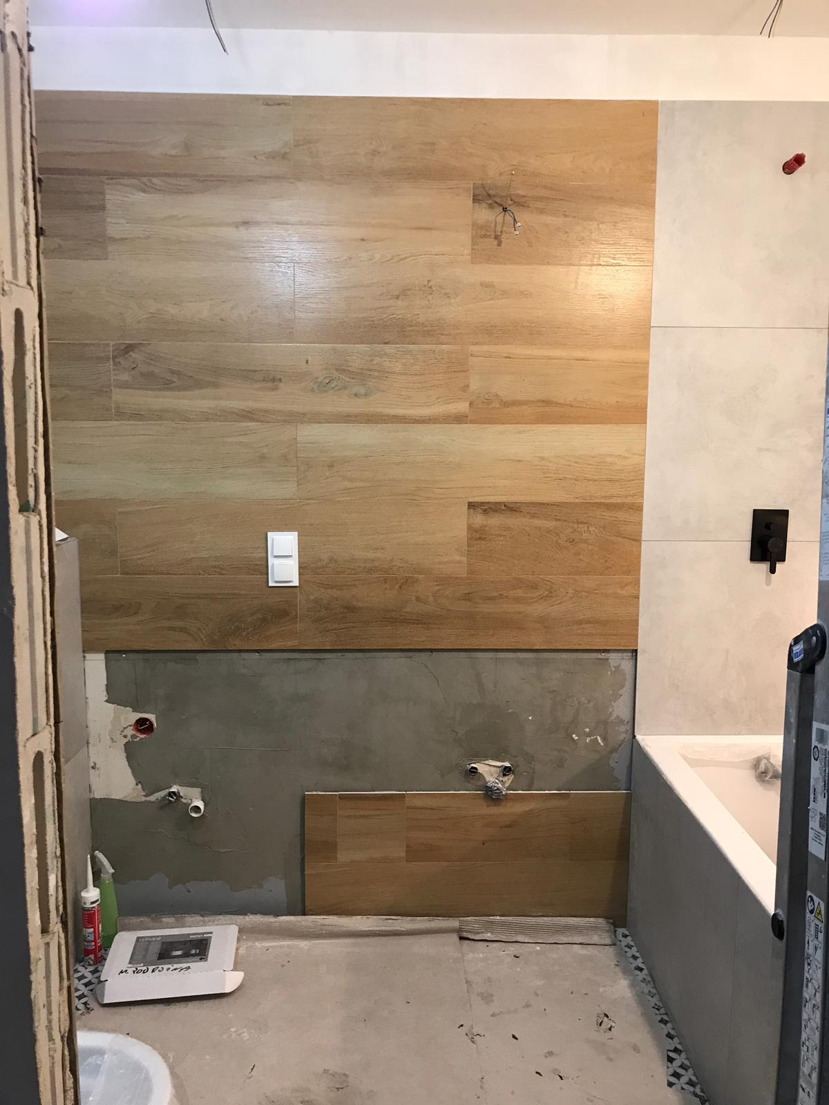 Łazienka w trakcie remontu: ściana wyłożona płytkami drewnopodobnymi w górnej części, poniżej surowy beton z wystającymi rurami, obok fragment ściany z jasnymi płytkami i czarną armaturą, widoczna...