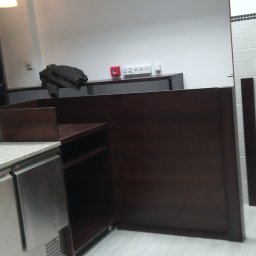 Alfa Producent mebli - Nowa zabudowa kuchenna z ciemnego drewna i metalowymi szafkami chłodniczymi, widoczna część osoby w czarnej koszulce, w tle białe płytki i sprzęt budowlany.