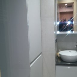 Alfa Producent mebli - Nowoczesna łazienka z wysoką, zabudowaną szafą w odcieniach szarości, umywalką nablatową i lustrem z odbiciem osób.