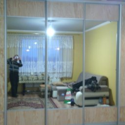 Alfa Producent mebli - Trzydrzwiowa szafa przesuwna z lustrami, odbicie pokoju dziennego z widoczną osobą robiącą zdjęcie.