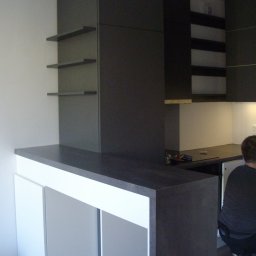 Alfa Producent mebli - Nowoczesna kuchnia z ciemnymi szafkami i białym barem, widoczne narzędzia na blacie i osoba w tle.