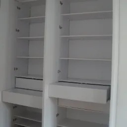 Wnętrze białej, otwartej szafy z półkami i wysuwanymi szufladami, widoczne zawiasy i drewniana podłoga.