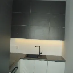 Nowoczesna kuchnia: ciemne, kwadratowe szafki wiszące z podświetleniem LED nad białymi szafkami dolnymi i ciemnym blatem ze zintegrowanym zlewem i czarną baterią.