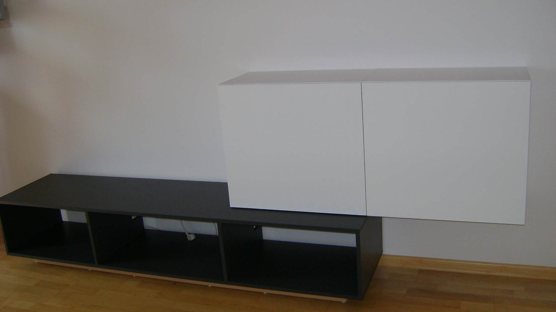 Nowoczesny zestaw mebli: ciemnoszara szafka RTV z otwartymi półkami i biała szafka wisząca na ścianie, na tle parkietu.