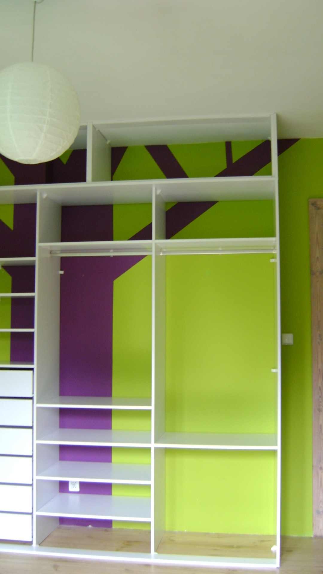 Biała szafa wnękowa bez drzwi z półkami i drążkami na ubrania, na tle ściany pomalowanej w geometryczne wzory w kolorach zielonym i fioletowym, z wiszącą papierową kulą-lampą.