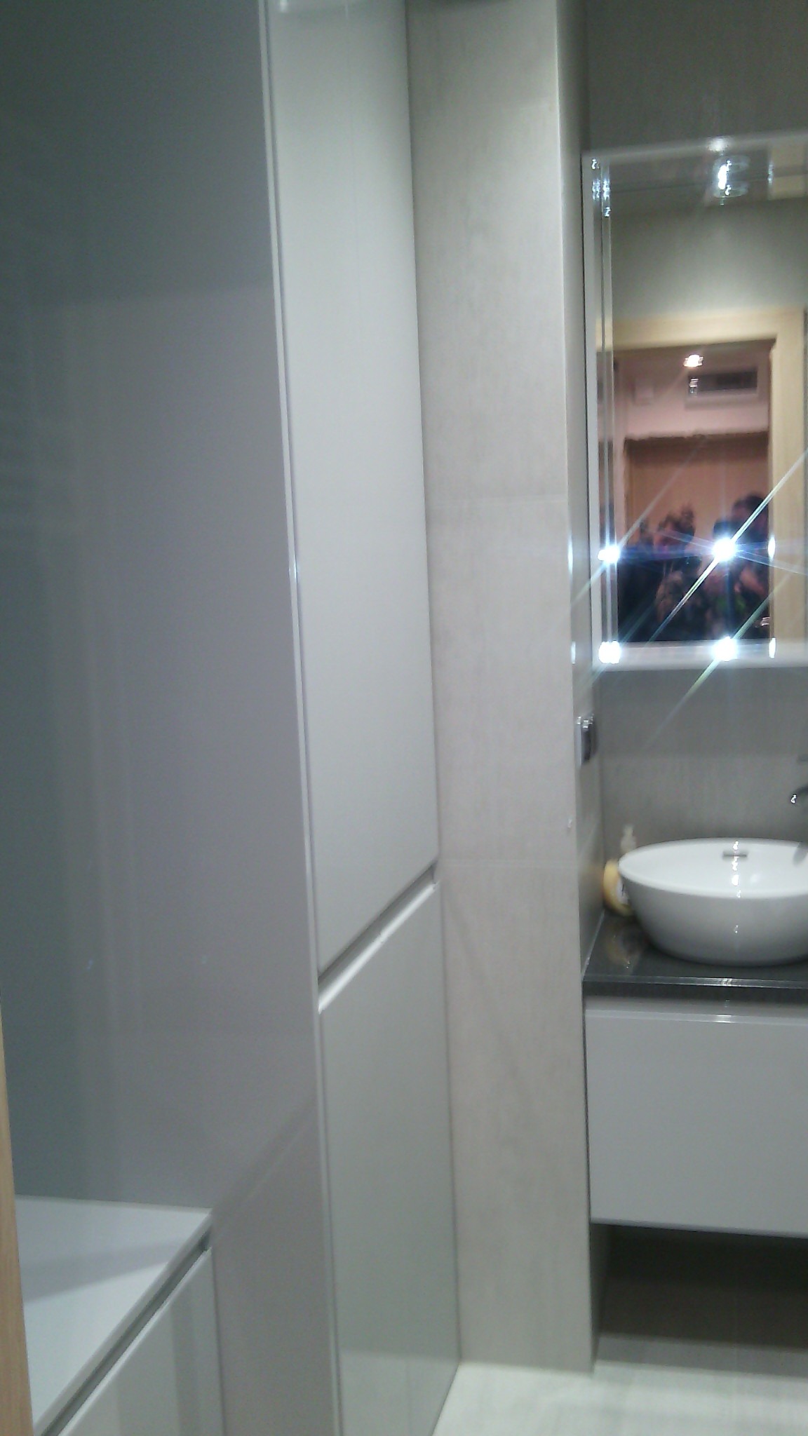 Nowoczesna łazienka z wysoką, zabudowaną szafą w odcieniach szarości, umywalką nablatową i lustrem z odbiciem osób.