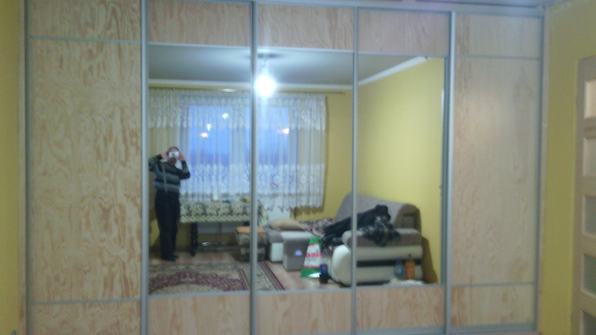 Trzydrzwiowa szafa przesuwna z lustrami, odbicie pokoju dziennego z widoczną osobą robiącą zdjęcie.