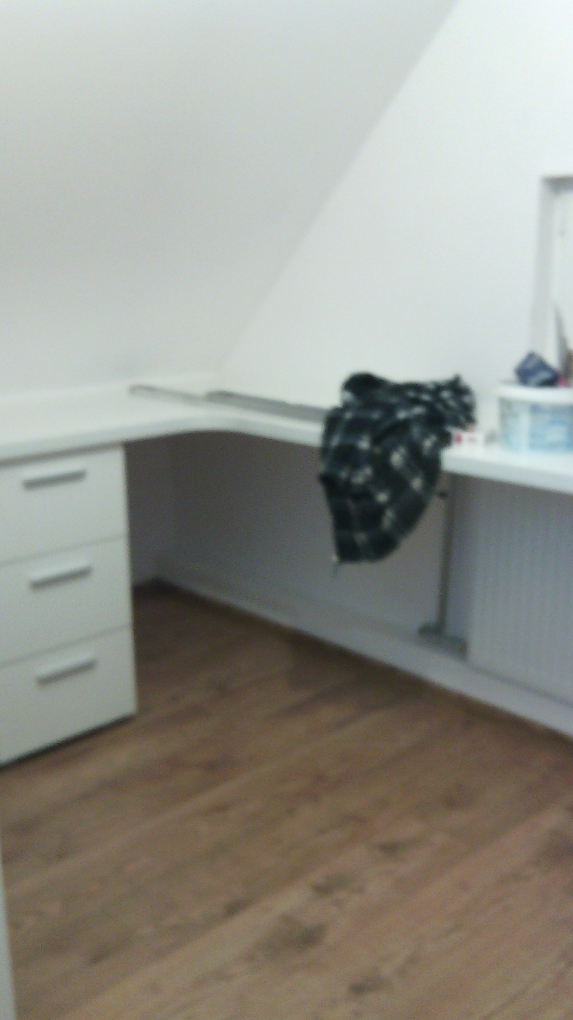 Białe biurko z szufladami i blatem w rogu pokoju na poddaszu, z drewnianą podłogą, częściowo zasłonięte czarną tkaniną w białe wzory, ujęcie nieostre.