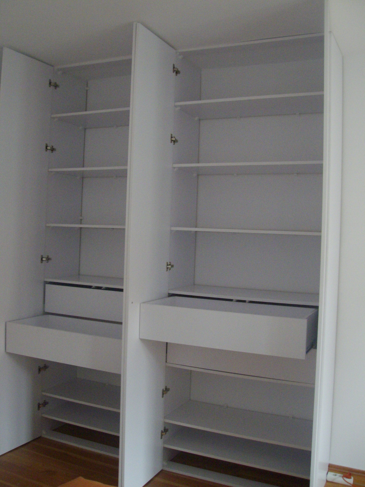 Wnętrze białej, otwartej szafy z półkami i wysuwanymi szufladami, widoczne zawiasy i drewniana podłoga.