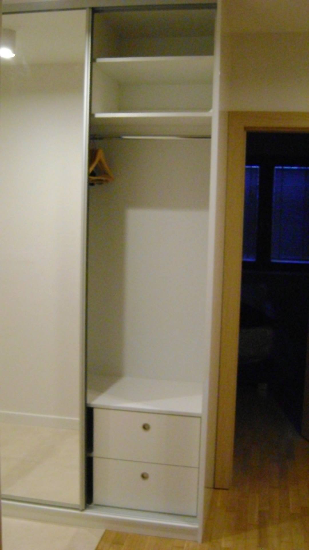 Biała szafa wnękowa z przesuwnymi drzwiami lustrzanymi, widoczne półki, drążek na ubrania z jednym wieszakiem i dwie szuflady z okrągłymi gałkami, na podłodze parkiet.