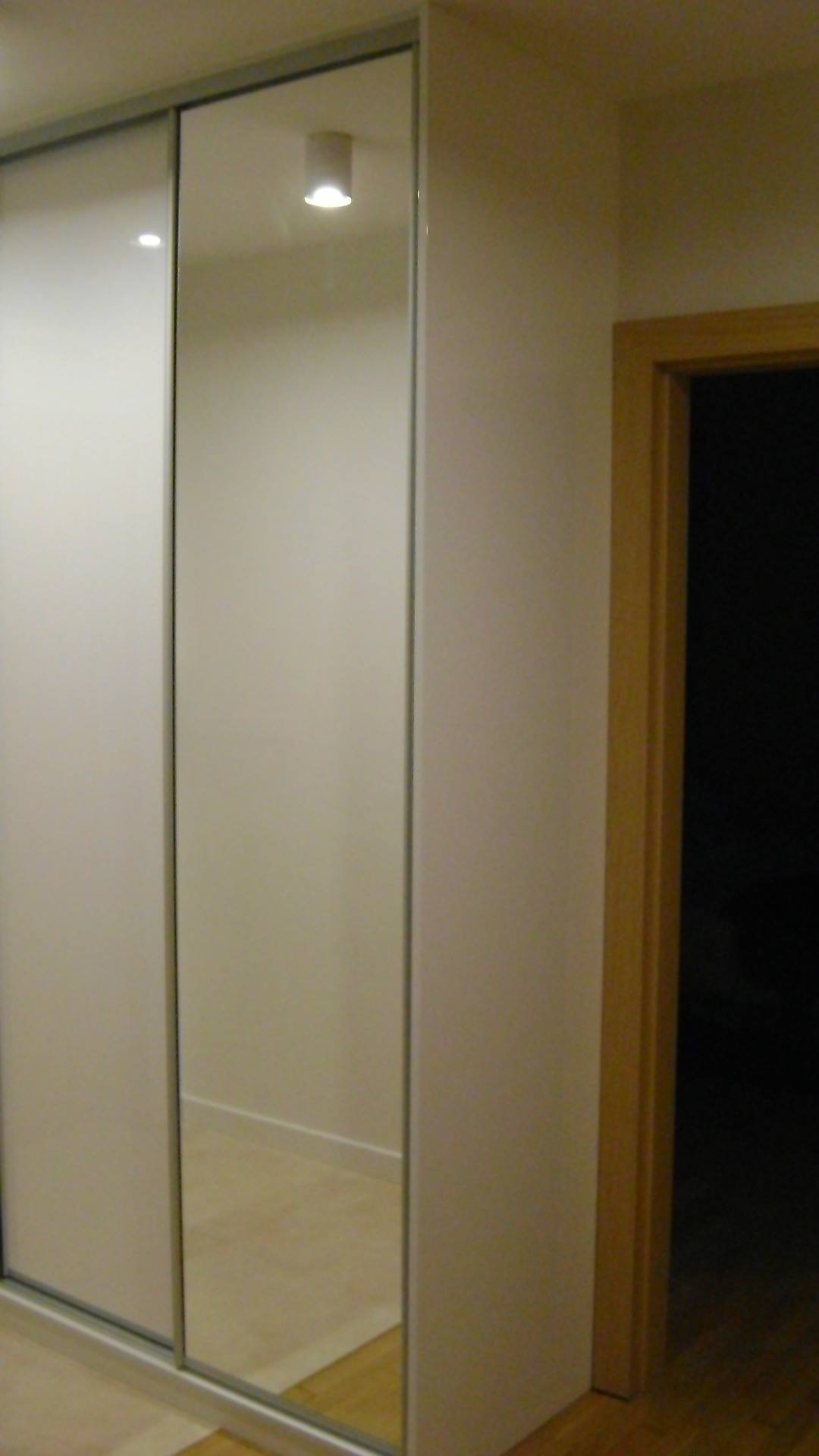 Biała szafa przesuwna z lustrzanymi drzwiami w korytarzu, widoczne odbicie podłogi i sufitu z lampą.